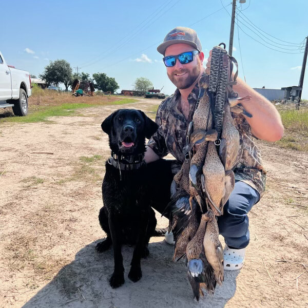 Hunter kneeling with a black Labrador and a strap of teal ducks on a sunny day